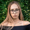 Young woman with long blonde hair and glasses against green foliage.