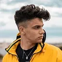 Side profile of a young man with voluminous dark hair and a yellow jacket.